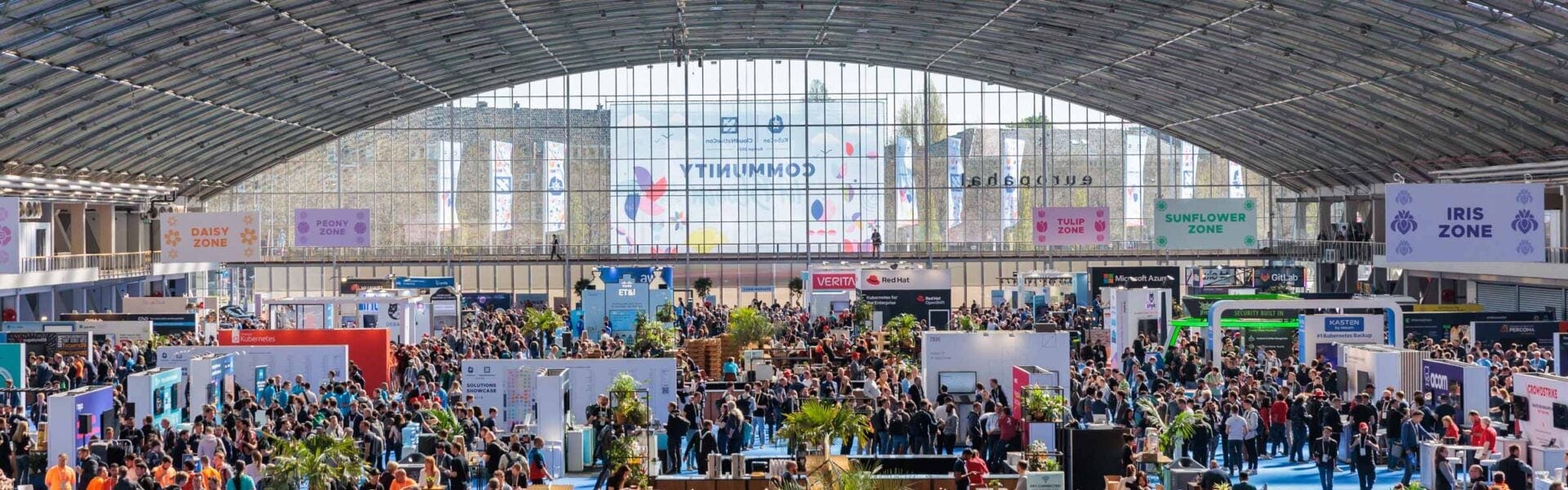 Wide shot of a busy event expo hall with attendees walking between booths. The floor is organized into color-coded zones labeled with flower names like "Tulip Zone" and "Iris Zone." Each booth features branding and displays, and the atmosphere is lively and well-lit.