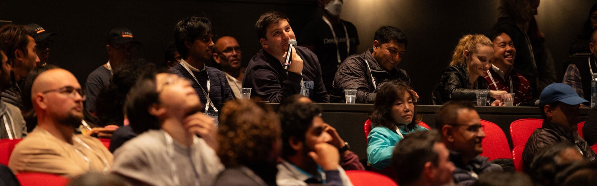 Audience members seated at a conference, with several people smiling and laughing. A man in the background smiles up at the stage while holding a microphone.