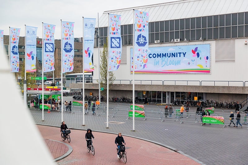 Exterior of RAI Amsterdam with "Community in Bloom" signage and colorful event flags, as people bike past the entrance.