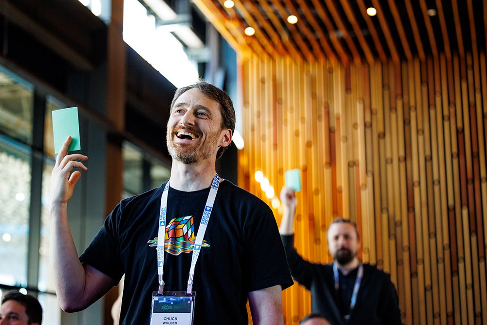 Attendees smiling and holding up green cards during an interactive onsite activity at a conference. A man in the foreground wearing a Rubik’s Cube t-shirt and conference badge is laughing and engaged.