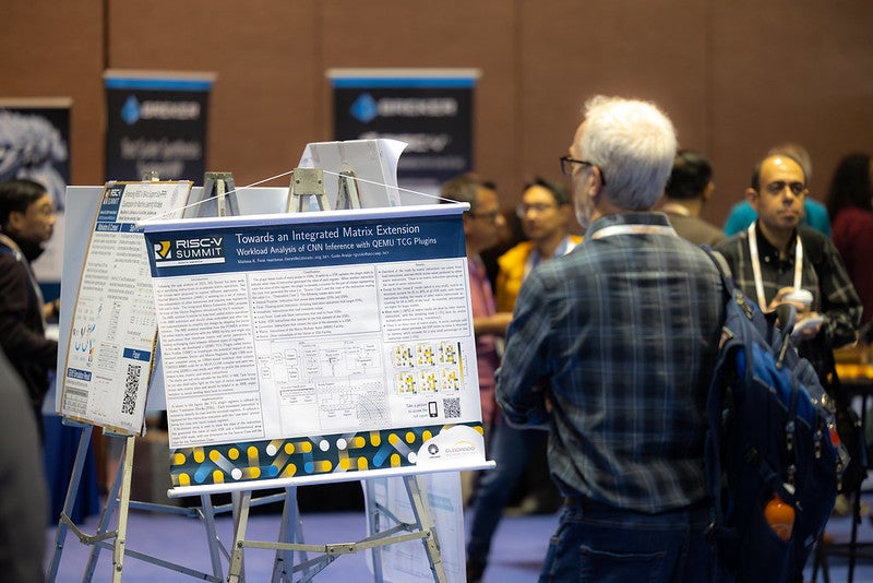 A man standing in front of a poster, in a room full of people.