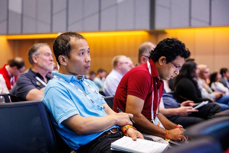 Two men in a room full of attendees listening to a speaker.