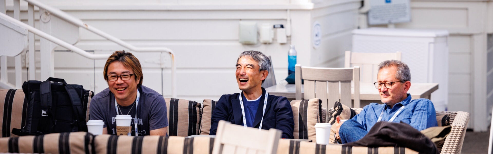 Three men sit on an outdoor couch, laughing and chatting together.