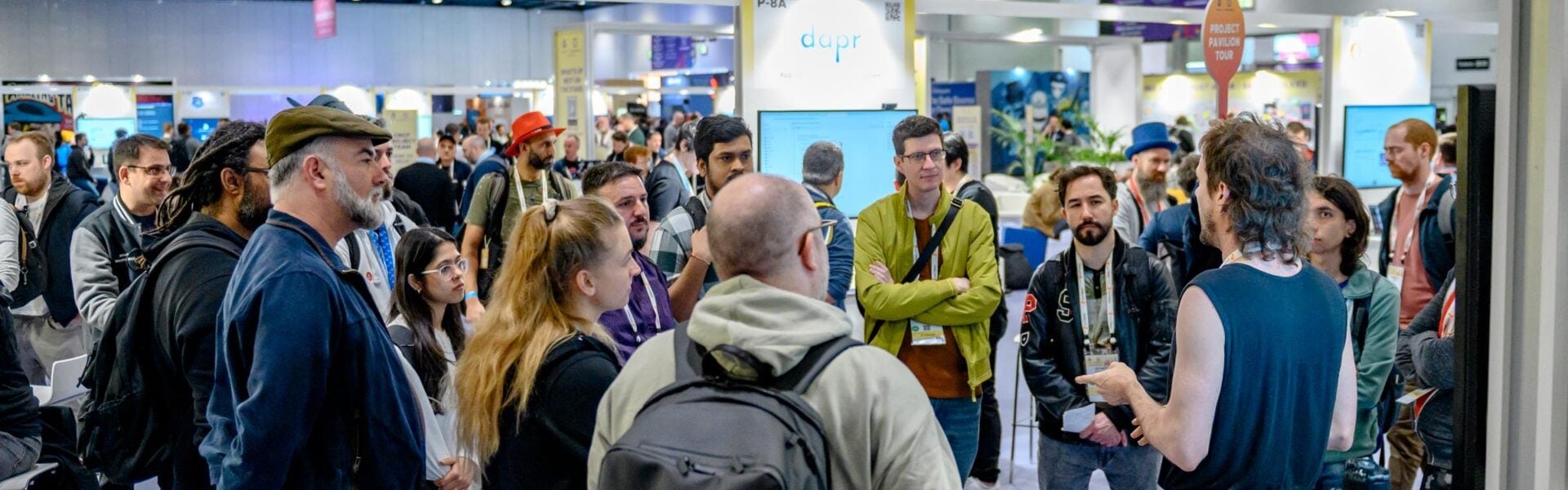 A man in a blue shirt speaks to a group gathered in a circle during a tour of the Project Pavilion. The room is busy with event attendees and booths, and a person in the background holds an orange “Project Pavilion Tour” sign while wearing a blue top hat.