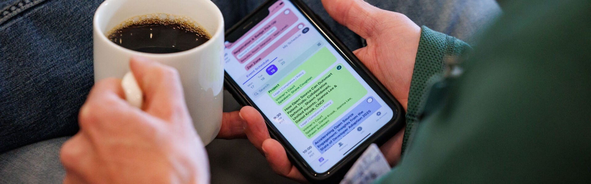 Close-up of a person holding a cup of coffee in one hand and a phone displaying an event schedule in the other.