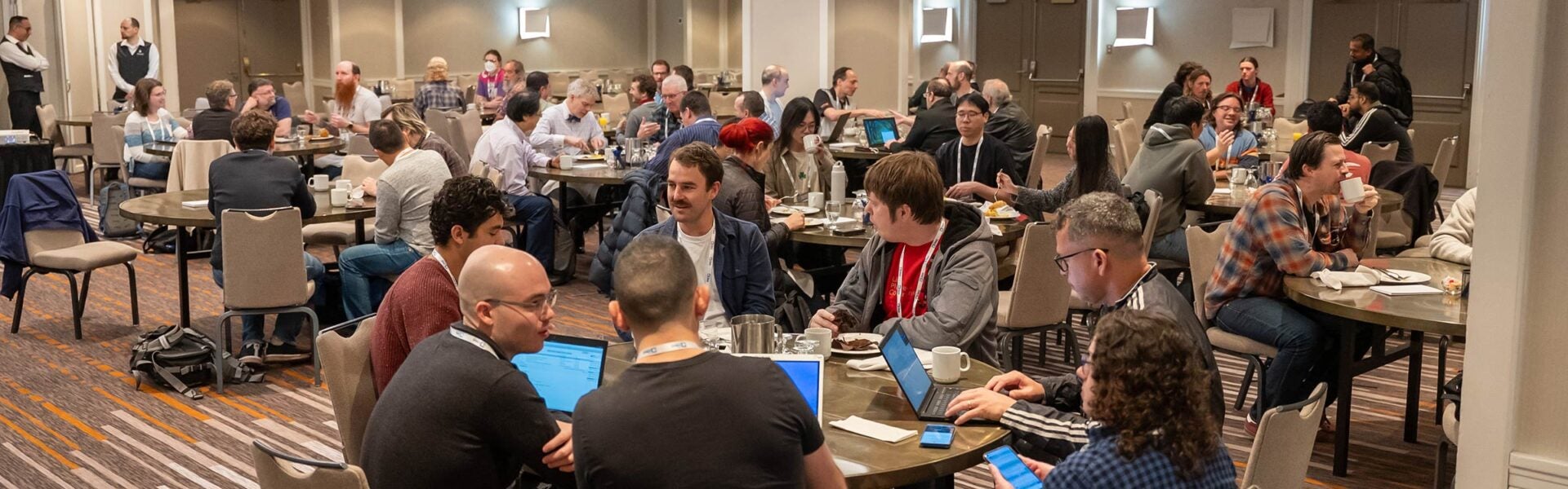 Attendees seated around round tables working on their computers, having food & coffee & visiting with each other.