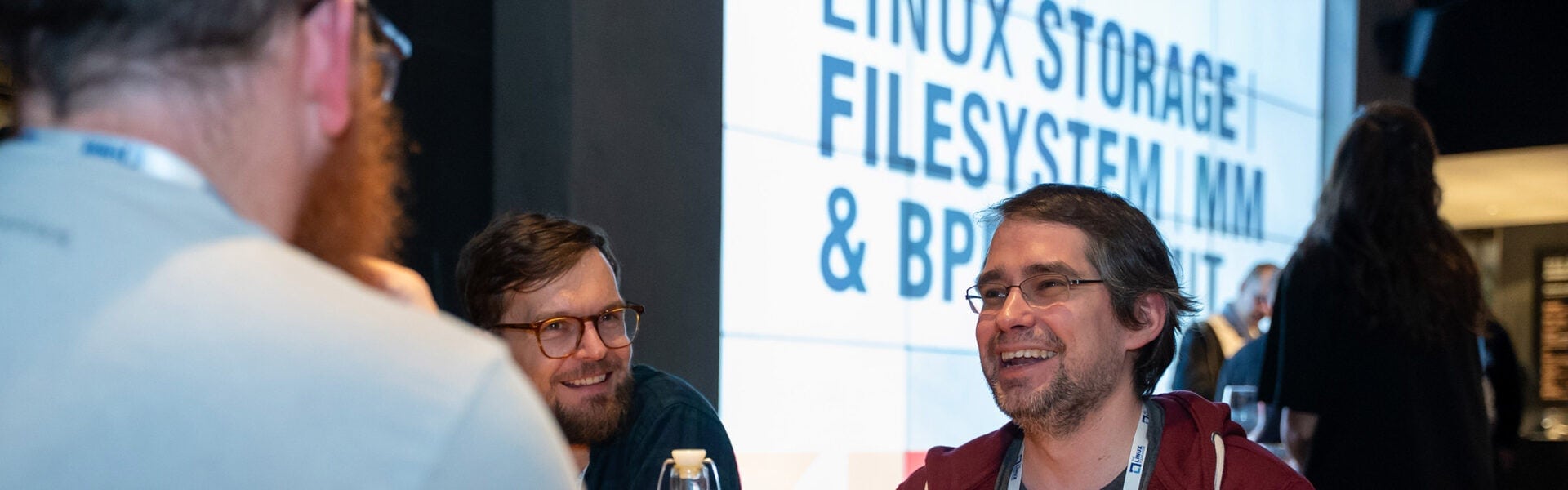 Three men talking and laughing in front of a sign that says Linux Storage | Filesystem | MM & BPF Summit.