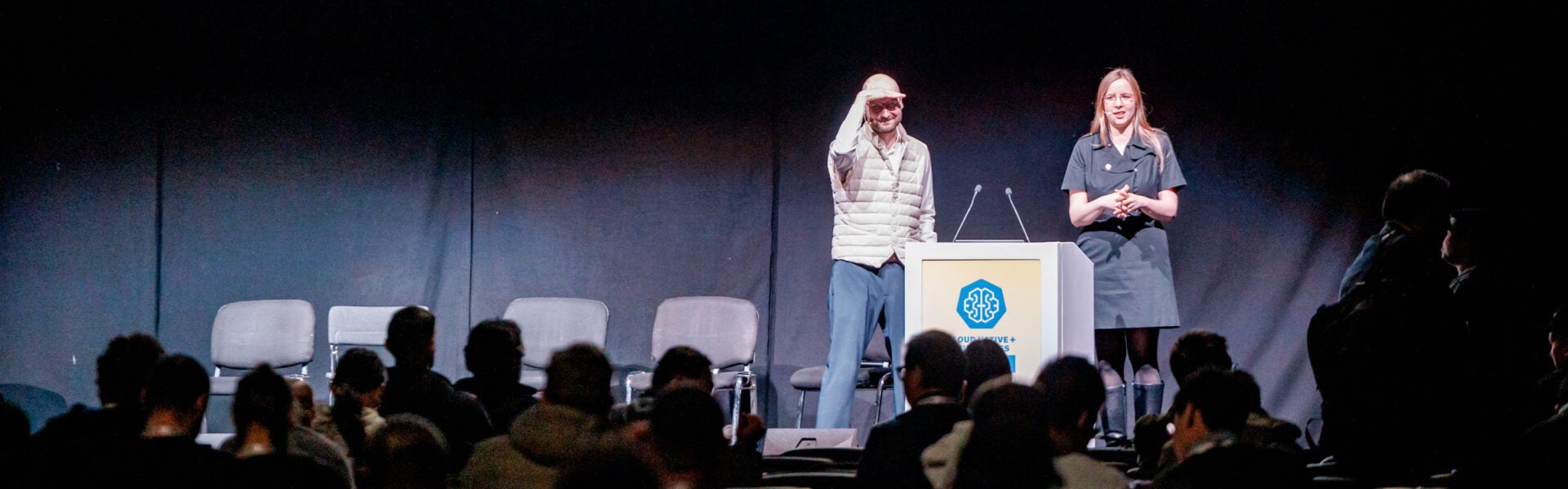 Two speakers on stage, one of them is shielding his eyes from the lights to see the audience.