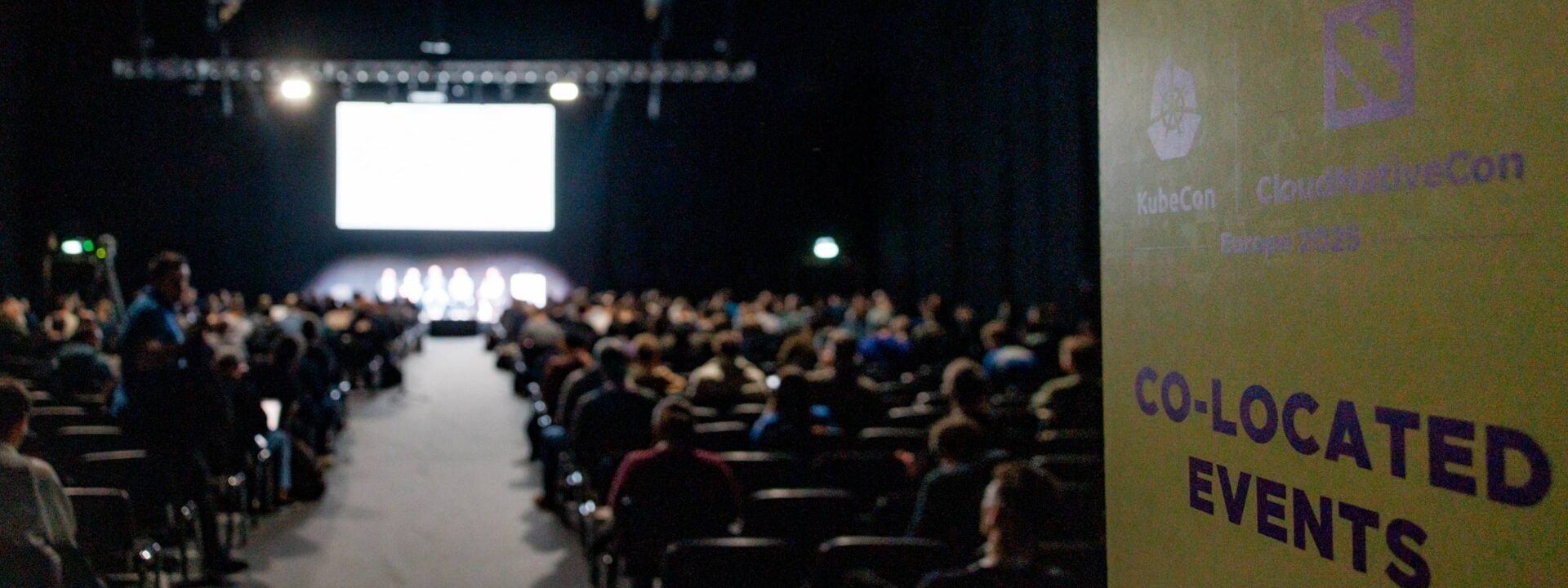 An auditorium filled with attendees with 5 speakers on stage. In the foreground is a sign that says "KubeCon + CloudNativeCon Co-located Events."