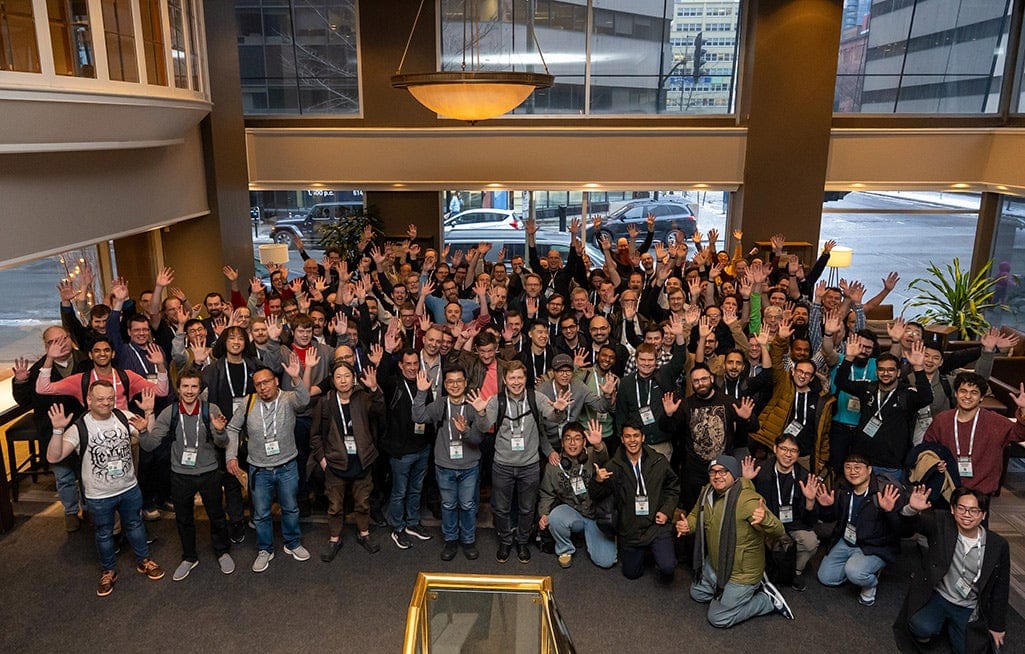 A large group of event attendees gathered indoors, smiling and raising their hands for a group photo.