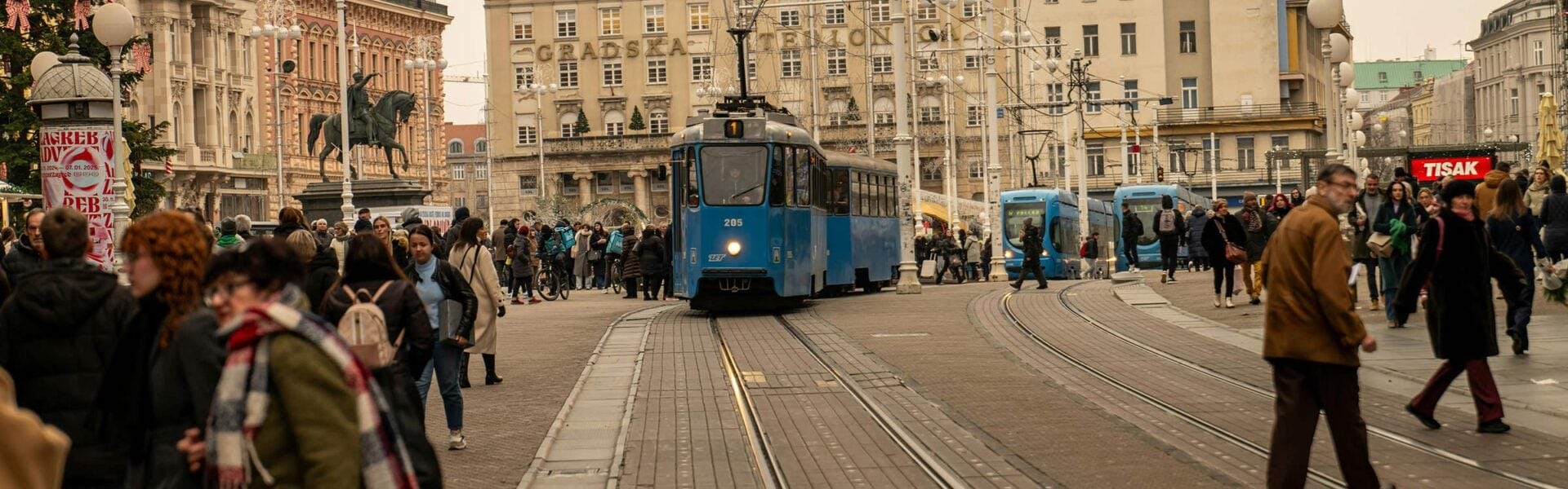 Three blue trams on tracks running on a street in Zagreb with pedestrians on either side.