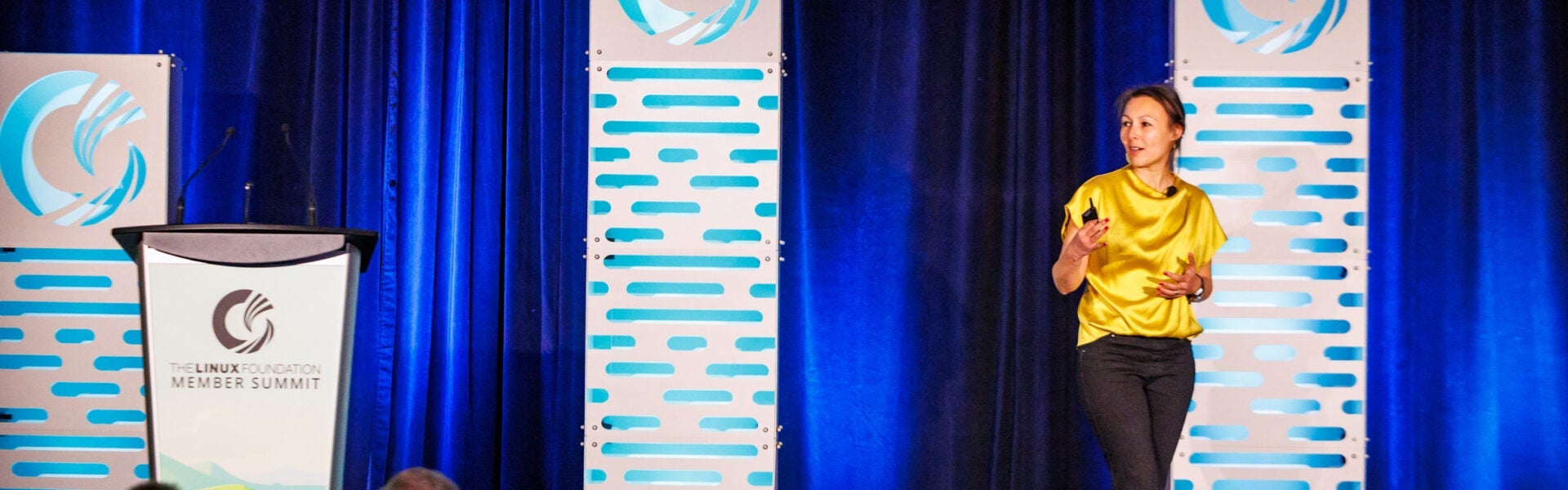 A woman wearing a yellow blouse, speaking on a stage with blue curtains.
