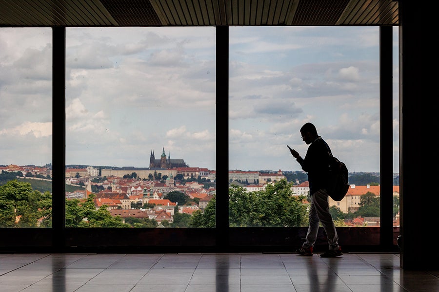 A person with a backpack checks their phone while standing inside the Prague event venue, with large windows framing a panoramic view of the city and Prague Castle in the distance.