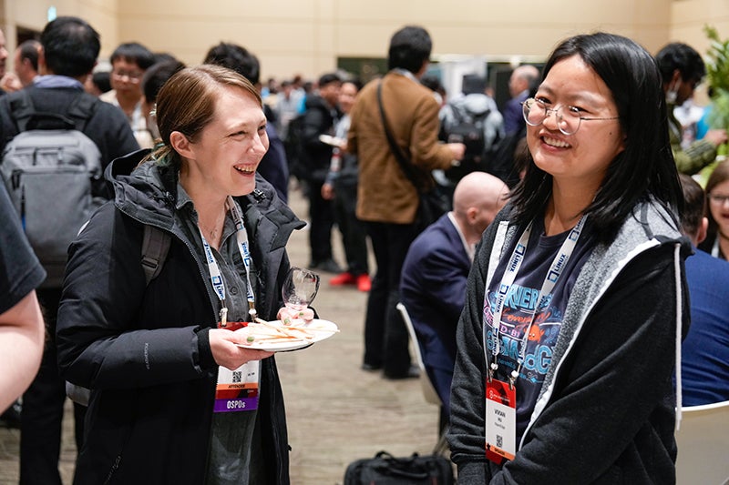 Two conference attendees smile and chat during a networking reception, one holding a small plate of food and a drink, with other participants mingling in the background.