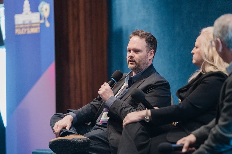A man sitting on staging with other speakers, holding a microphone.