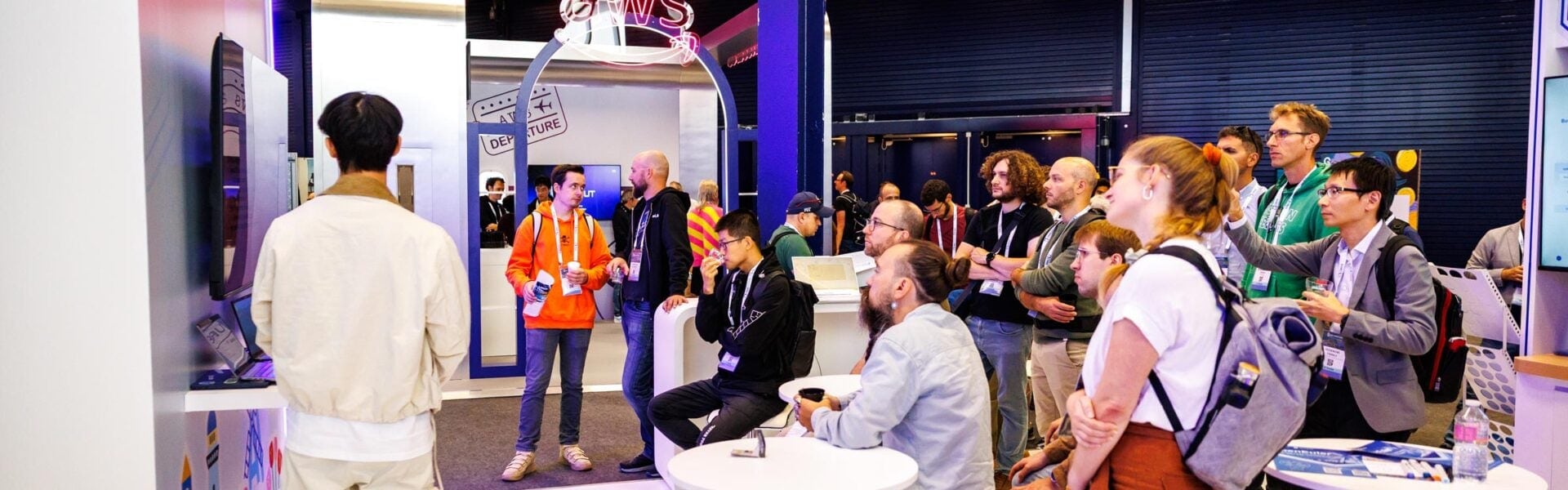 Attendees gather around a booth, watching a presentation on a screen at a conference.