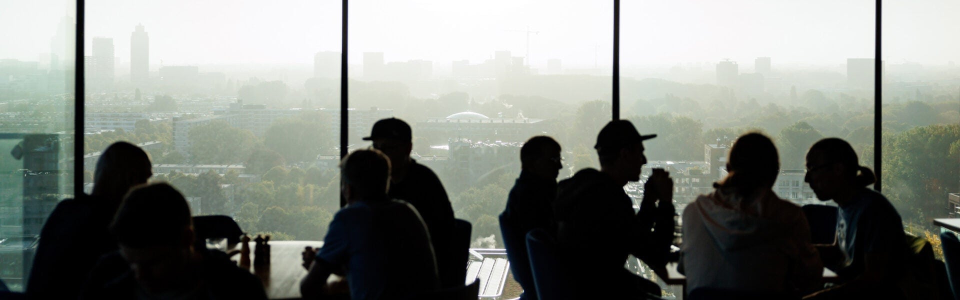 Attendees sit at tables near tall windows, silhouetted against a hazy city skyline.