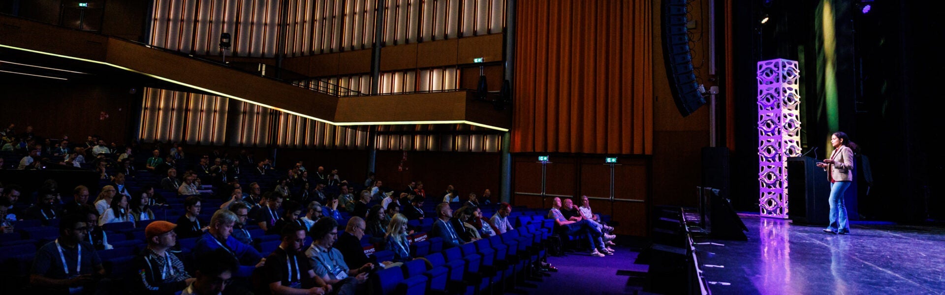 A presenter speaks on stage in front of a large audience in a theater-style setting.