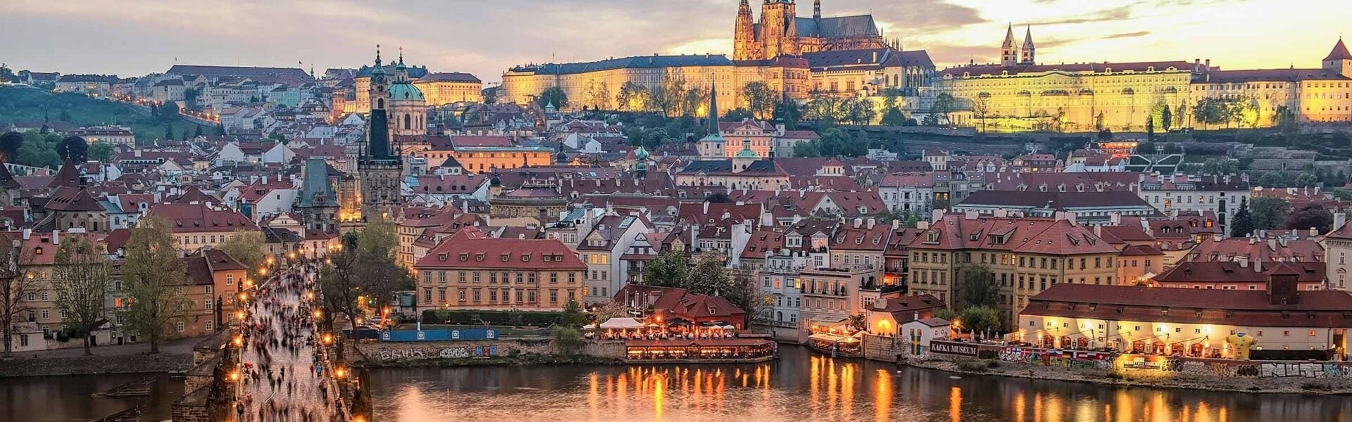 View of Prague from the water, with people walking along a bridge and buildings along the shore.