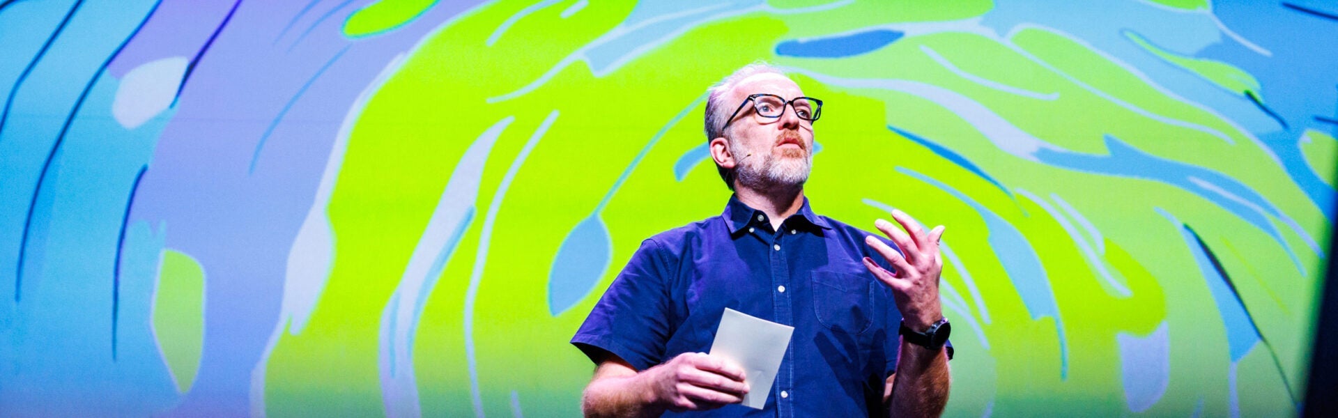 A speaker presents on stage at Open Source Summit, gesturing with one hand while holding notes in the other, with a bright abstract design projected behind them.
