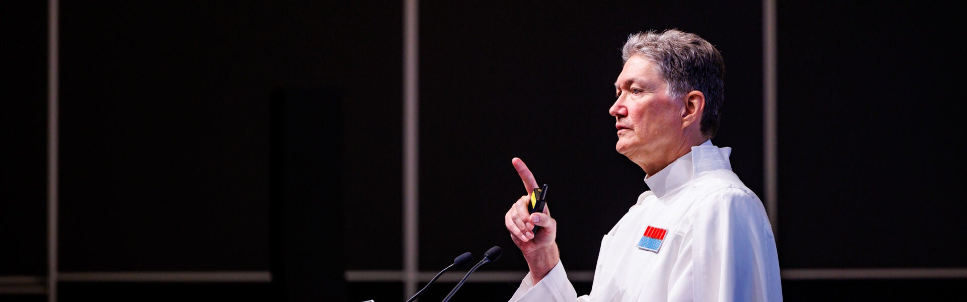 A man in a white coat standing in front of microphones with his index finger raised.