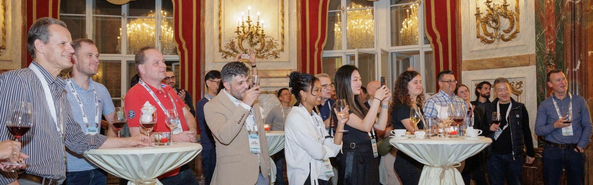 Attendees enjoy drinks and conversation in an elegant venue with chandeliers and ornate decor during an evening reception.