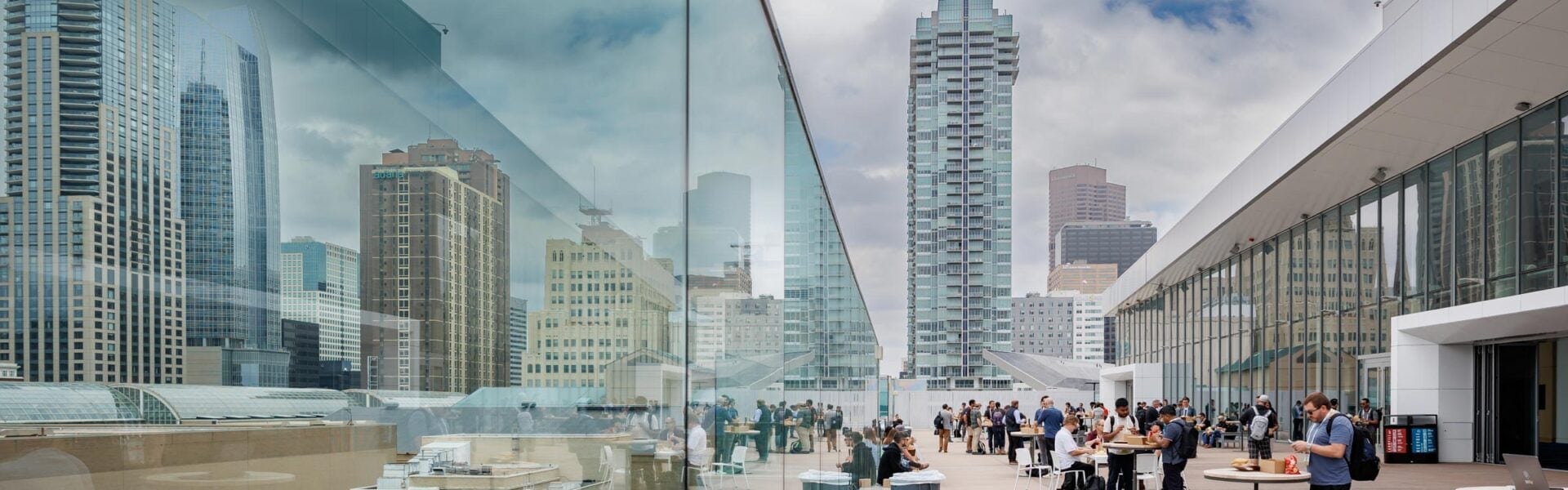 Attendees gather on a rooftop terrace with the city skyline reflected in floor-to-ceiling glass, chatting and eating at standing tables.