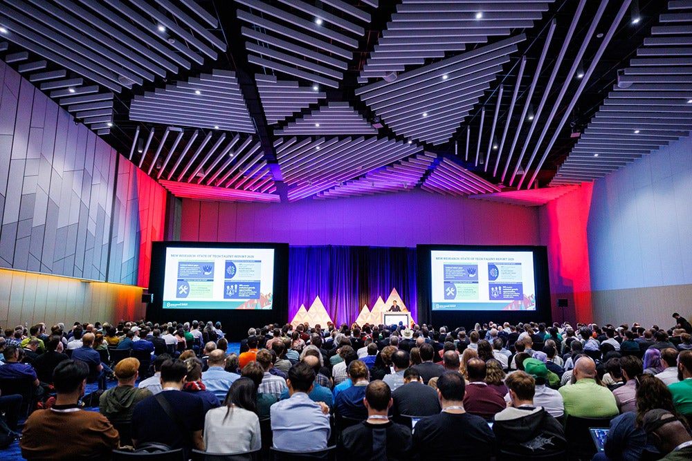A large audience seated in a modern conference hall watching a presentation on stage, with two large screens displaying slides and colorful lighting illuminating the ceiling.