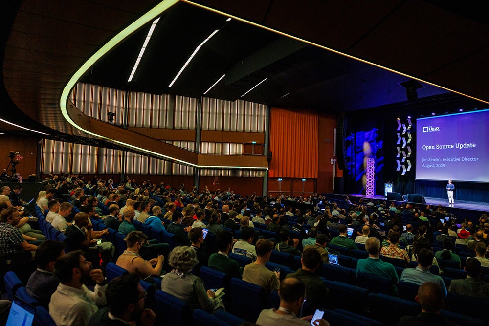 Audience seated in a large auditorium watching a keynote presentation with a slide reading “Open Source Update – Jim Zemlin, Executive Director, August 2025.