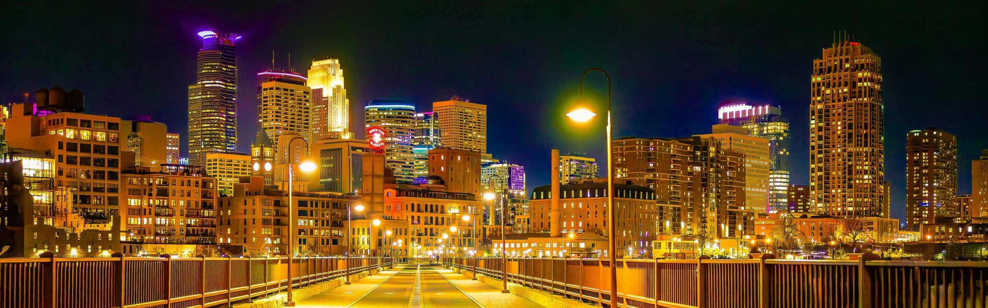 Nighttime view of the Minneapolis skyline from the Stone Arch Bridge, with glowing streetlights leading toward brightly lit skyscrapers under a dark sky.