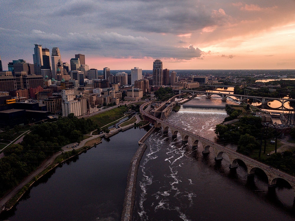 Aerial view of downtown Minneapolis at sunset, showing the Stone Arch Bridge, Mississippi River, and surrounding cityscape.