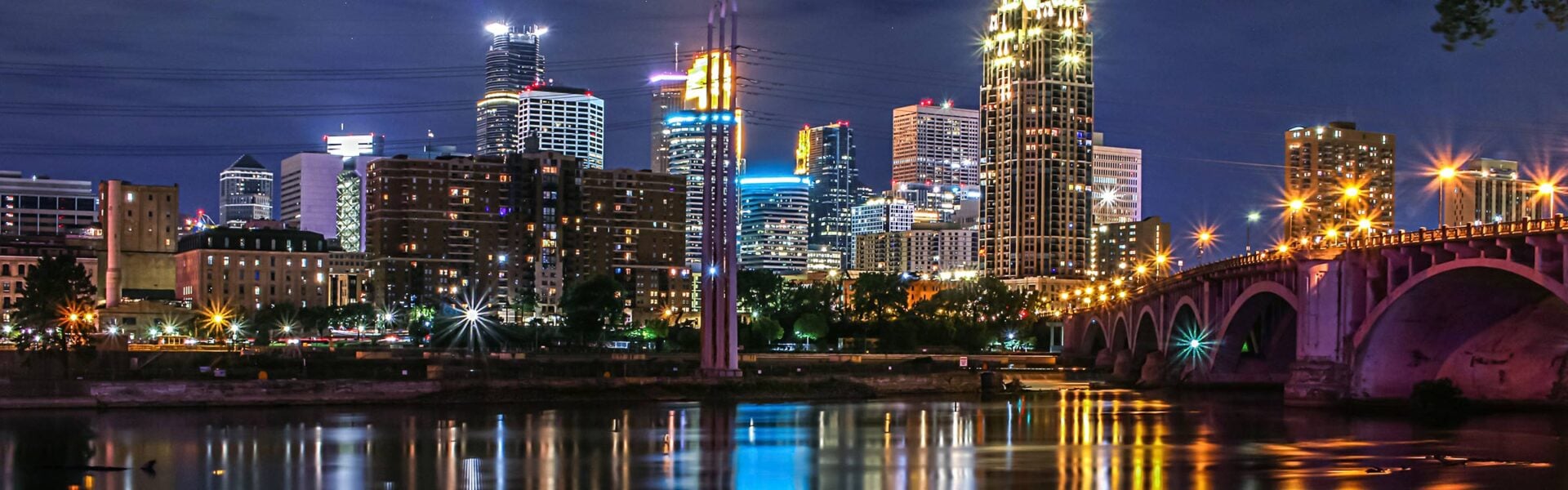 Minneapolis skyline illuminated at night, with bright city lights reflecting on the Mississippi River and the bridge crossing into downtown.