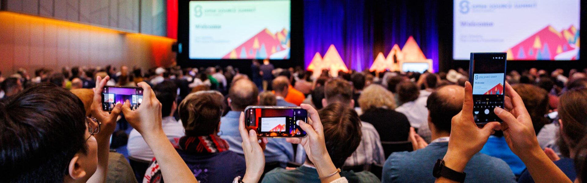 Audience members at the Open Source Summit raise their phones to capture photos of the keynote stage, where a welcome slide is displayed.