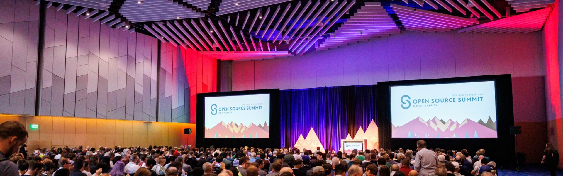 A packed keynote hall at the Open Source Summit, with two large screens showing the event logo and colorful mountain-themed graphics as attendees fill the room.
