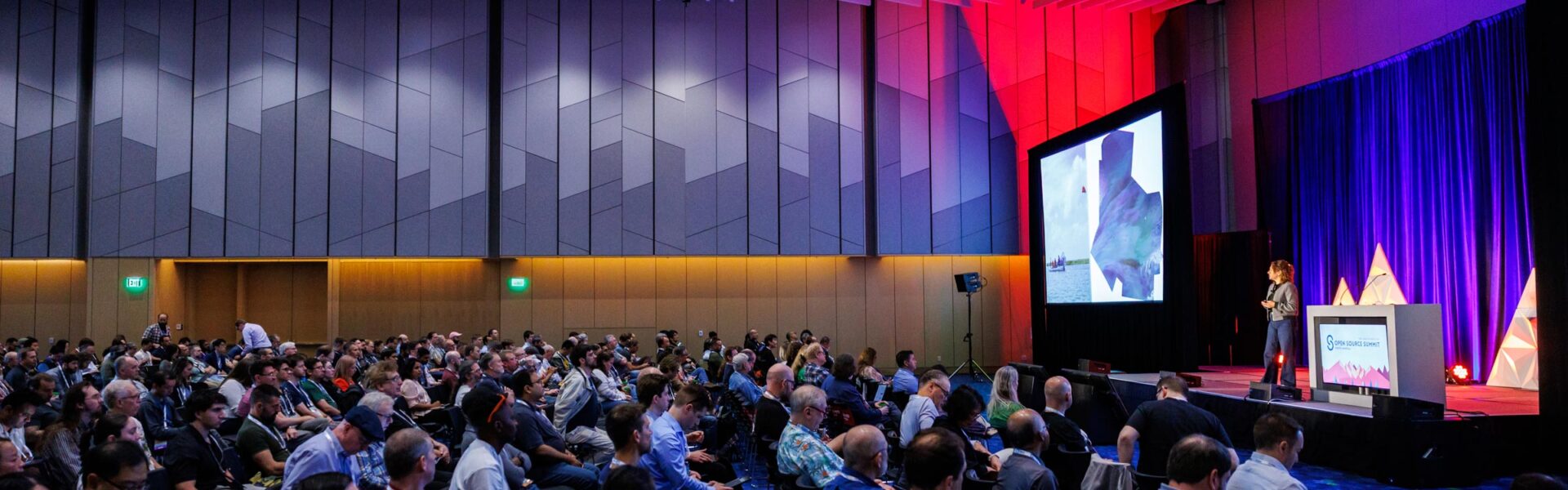 A speaker presents on stage at Open Source Summit, addressing a large seated audience in a conference hall illuminated with purple and red lighting.