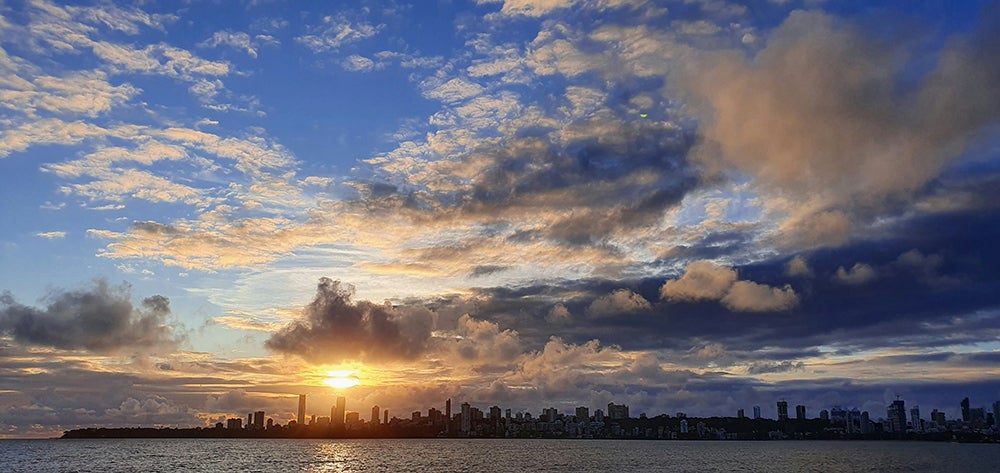 Sunset over Mumbai’s skyline seen from across the Arabian Sea, with golden light breaking through dramatic clouds above the cityscape.