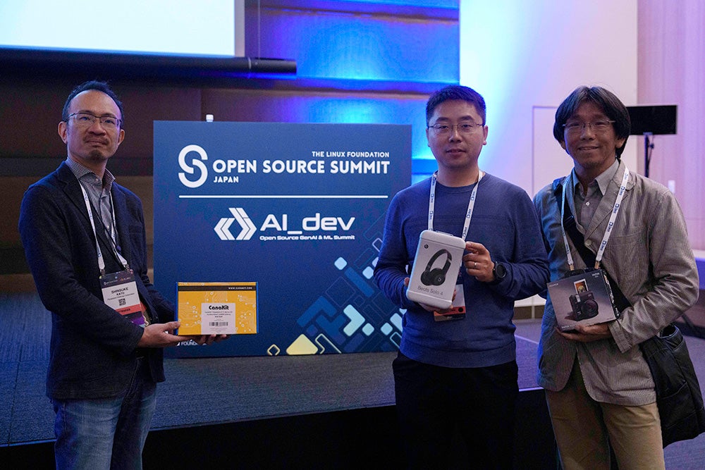 Three attendees stand on stage holding tech prizes in front of the “Open Source Summit Japan – AI_dev” sign.