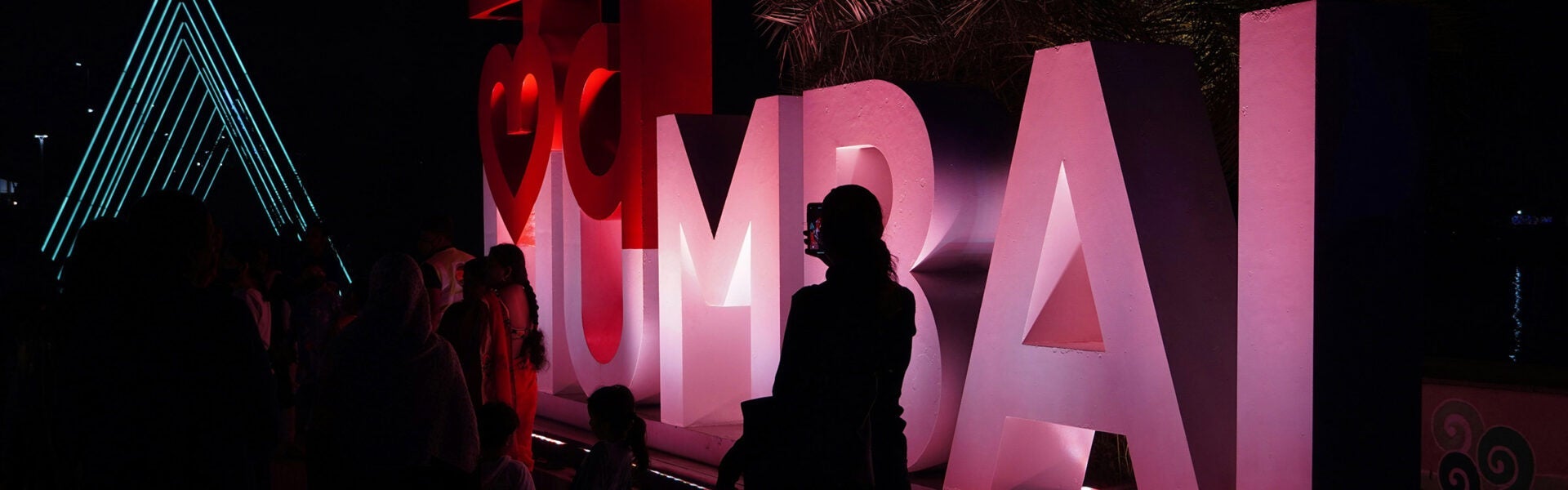 People silhouetted at night in front of an illuminated “I heart Mumbai” sign glowing pink and red, with neon blue light structures in the background.