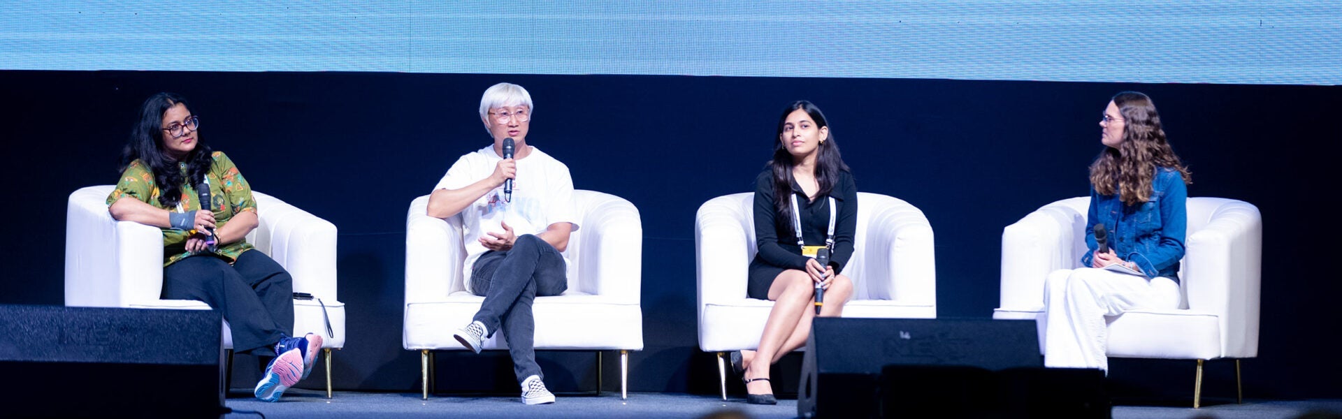 Four panelists seated on stage in white chairs during a conference discussion, each holding a microphone under stage lighting.