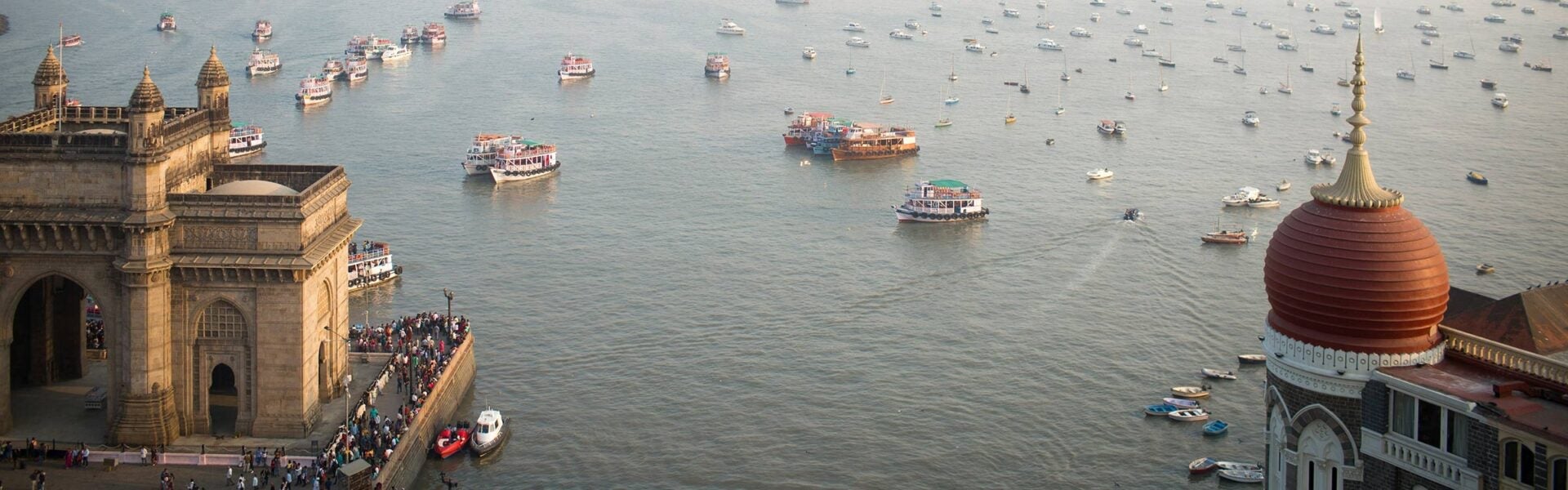 Aerial view of the Gateway of India and the Taj Mahal Palace Hotel in Mumbai, with boats and ferries scattered across the Arabian Sea.