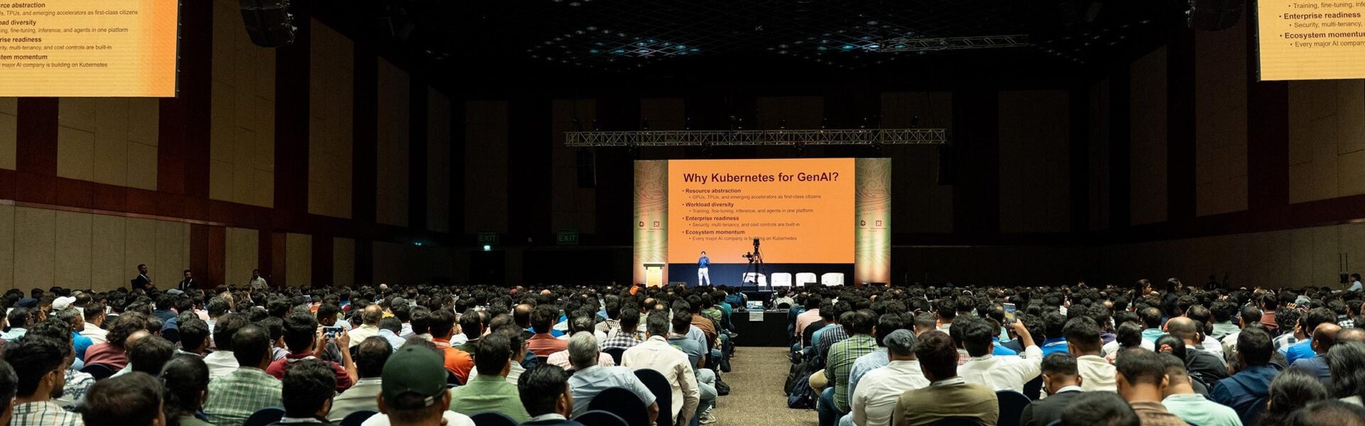 A large audience seated in a conference hall listening to a speaker presenting on stage with a screen displaying the topic “Why Kubernetes for GenAI?”