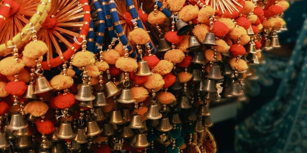 Colorful display of traditional decorative tassels and small metal bells, with bright pom-poms and woven rings in red, orange, yellow, and blue tones.