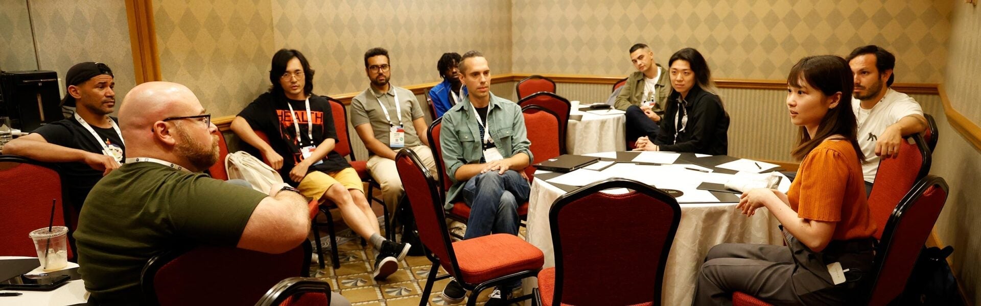 Small group of conference attendees sit in a breakout room, listening and discussing around round tables with notebooks and laptops.