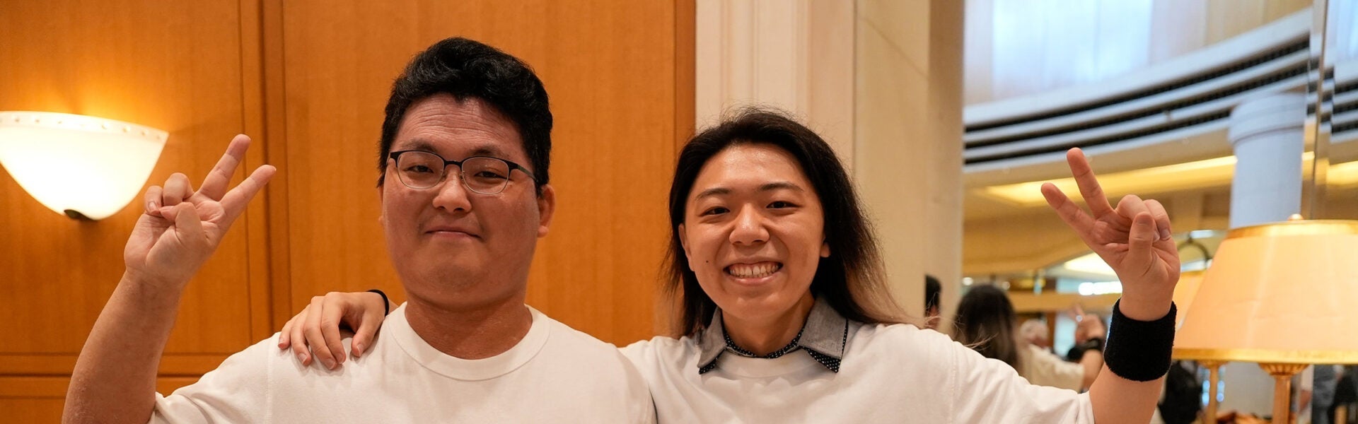 Two people smiling and posing indoors with peace signs, standing close together in a warmly lit conference venue.