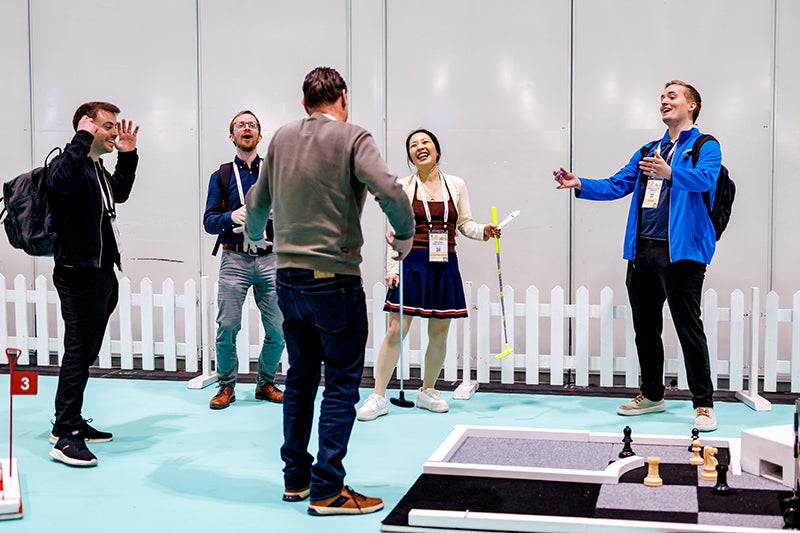 Five conference attendees laugh and interact around a mini-golf putting area and large chess board in an indoor event space.