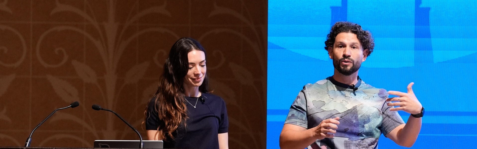 Two conference speakers on stage, one standing at a podium and the other gesturing while presenting in front of a blue screen.