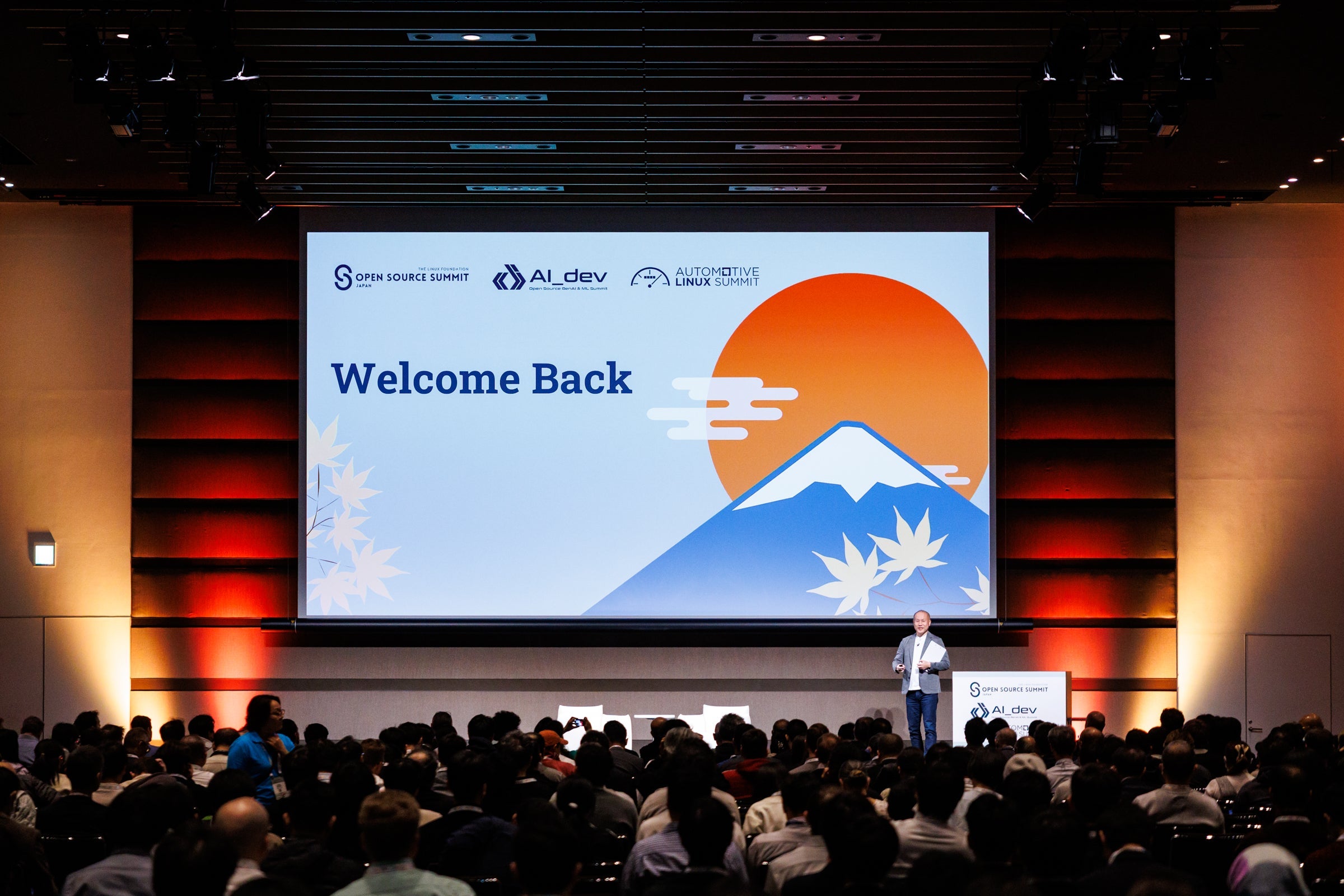 A crowded conference audience faces a stage with a “Welcome Back” screen and a speaker talking.