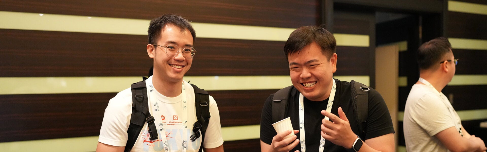 Two attendees smiling and chatting in a hallway at a conference, each wearing backpacks and lanyards.