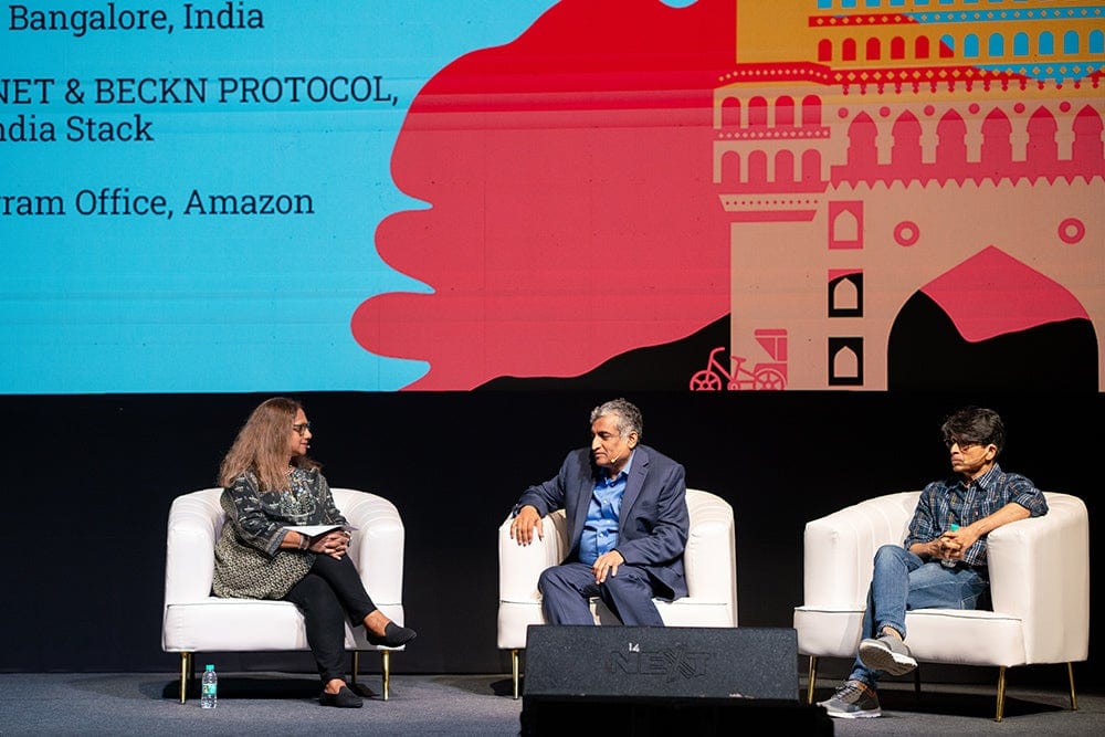 Three panelists sit in white armchairs on a conference stage, talking in front of a colorful slide backdrop.