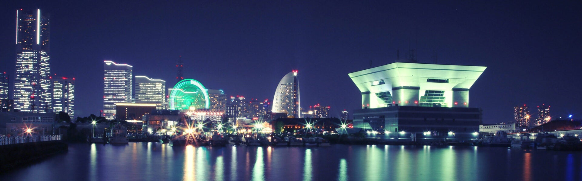A nighttime waterfront cityscape with illuminated skyscrapers, a glowing ferris wheel, and modern buildings reflecting across the water.